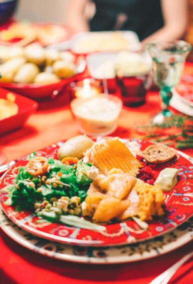 A huge spread of traditional Finnish Christmas food covers a long table with a red table cloth. The view is a close up of a plate full of food with water glasses and bowls of various foods arranged across the table.