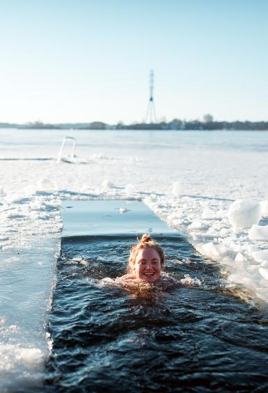 With the landscape of an ice covered sea dusted with snow stretching to the coast on the horizon, a smiling woman swims in a strip cut in the ice between trips to the sauna.