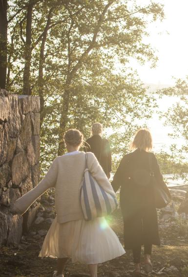 Three women carrying large bags are walking by a tall, dry-stone wall, past trees towards a rocky shoreline. The sun shine so brightly towards the camera that the water and view across it have whited out.