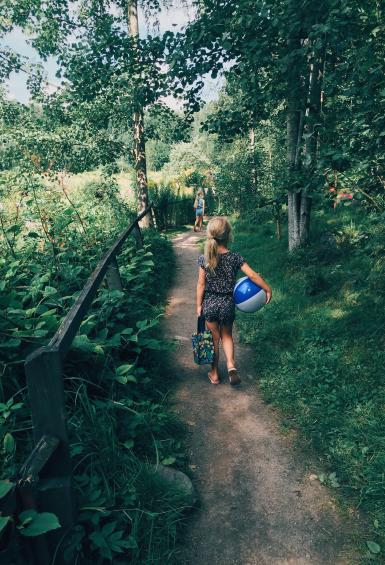 Facing away from the camera, a little girl carrying a beach ball under one arm and a bag in her opposite hand, walks down a trail that is surrounded by grass, bushes and trees.
