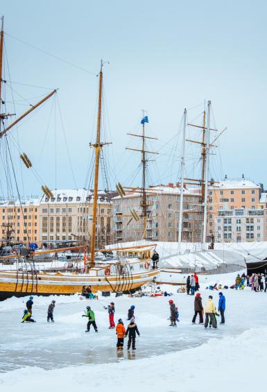 In a picture postcard like scene, people are skating on the ice at Halkolaituri dock in winter. A couple of sailing ships and colourful apartment blocks are in the background.