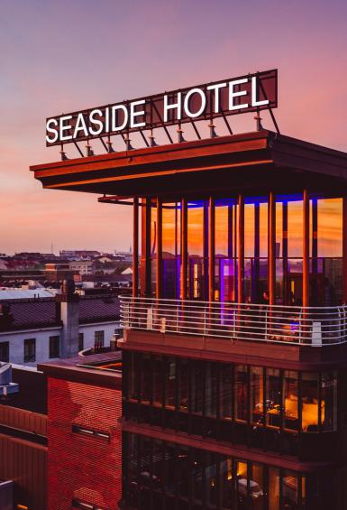 A view across the Helsinki rooftops stretches out behind the facade and rooftop terrace of Radisson Blu's Seaside Hotel under a pink and purple sky at sunset.