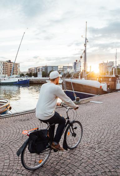 A man dressed in a white sweatshirt and cap, cycles away from the camera past docked boats, alongside west harbour as the sun sets. Apartment blocks are sat on the horizon on the edge of the harbour. 