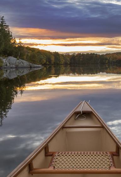 Sitting inside a canoe, with the front tip pointing forwards from the bottom of the photo, a sunset shines through from behind a cloudy sky, reflecting on a very calm lake surface. On the horizon and ahead to the left, a treeline stretches around the lake.