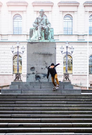 A skater performs a trick in front of a statue and the steps leading up to the Bank Of Finland behind them.