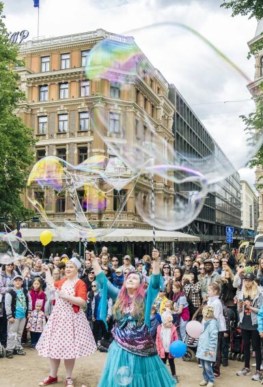 In Esplandi park, a large crowd of people on Helsinki Day watch performance artists blow giant bubbles. Hotel Kämp can be seen in the background.