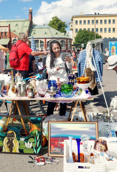 It's a sunny day and people are browsing through a variety of items at a Hietalahti Flea Market stall. Hietalahti Market Hall cann be seen in the background.