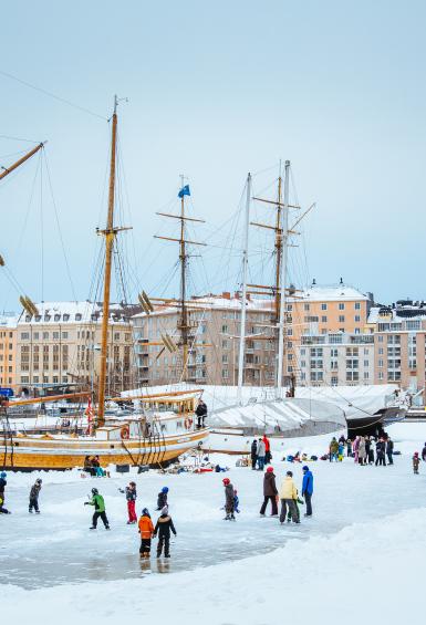 In a picture postcard like scene, people are skating on the ice at Halkolaituri dock in winter. A couple of sailing ships and colourful apartment blocks are in the background.