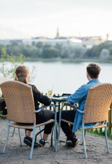 A couple sit together at Sinisen Huvilan Kahvila's outdoor terrace, looking out over Töölönlahti bay on a sunny day.