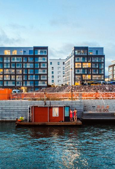 A small, red sauna ferry in the Kalasatama district during Helsinki Sauna Day, moored against a breeze block wall, apartments above it in the background.