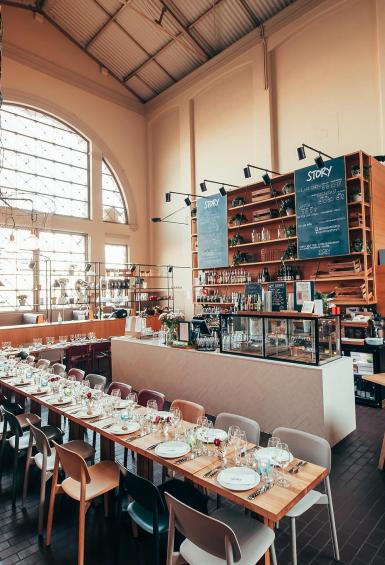Story restaurant inside Old Market Hall, a tall, open space with large windows towards the left and the main market leading away down a corridor to the right of the restaurant's main counter. Behind the counter are a set of tall, wide shelves whereas in front of the counter, a long table has been set with dozens of chairs for a private event.