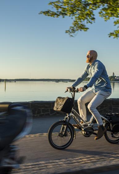 A man is cycling to the left of the photo on the path running by the Kaivopuisto shore, where the sea beyond is flat calm and the sun is shining.