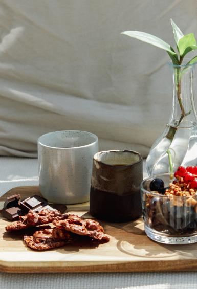 Sitting on a wooden serving board having been neatly arranged are a coffee mug, coffee in a small jug, a few cookies, some squares of chocolate, a berry and granola desert in a small glass tumbler and a fluted vase holding a small green plant.