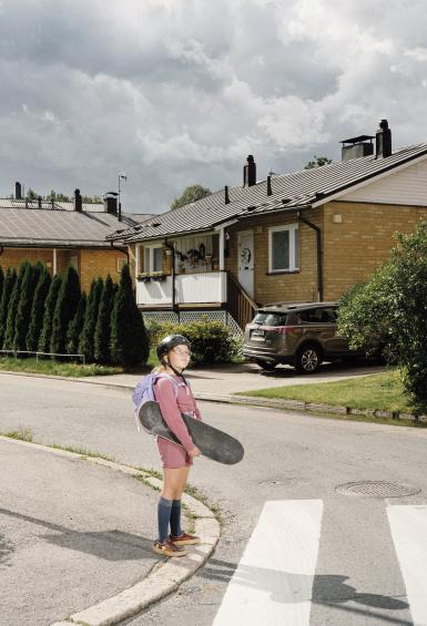 Selma is in her local suburban area, houses in the background, standing at the edge of a street crossing holding her skateboard under her arm.