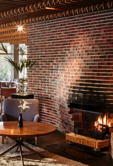 On the right, surrounded by a brick wall facade, is the fireplace at Hotel Rantapuisto's restaurant. Immediately on the left are a table and 3 armchairs, in the background are dozens of tables set for service in the restaurant. 