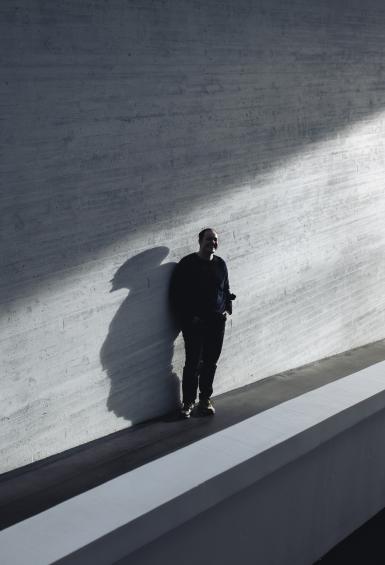 Jamie MacDonald standing in front of a grey wall along a narrow walkway in the mid-ground of the photo, a stream of sunlight shining down on him from the right.
