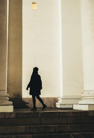 A silhouetted figure walking between a pair of large columns of a white stone neo-classical building, slate grey steps descending in the very bottom of the photo.