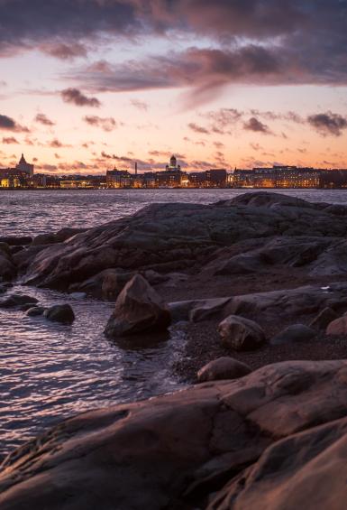 A view of Helsinki from a rocky shoreline during dusk with a pink sky. Helsinki Cathedral can be seen on top of the cityscape on the coastline in the distance. 