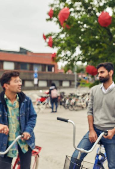 Three people are standing in front of Aalto University, a woman in the foreground looking straight at the camera, while 2 men mounted on bikes are chatting behind her. Behind them all are bike racks, a tree in full bloom dotted with red umbrellas, and the Aalto University buildings.