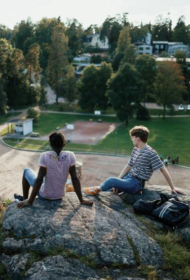 Two young guys are sitting on a rock overlooking a park surrounded by trees, enjoying the summer sun