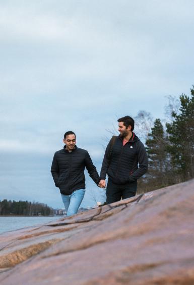 A male couple are walking across reddish rocks by the shore on an overcast day in Helsinki.