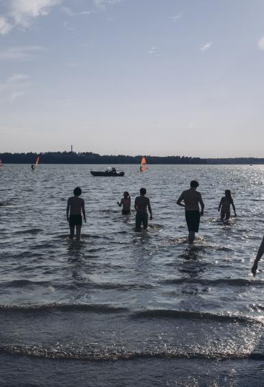 On a warm summer's day, sun glaring off the sea, a small group of people wade out into the water from the beach at Kasinonpuisto. The Servinniemi shoreline can be seen in the distance.