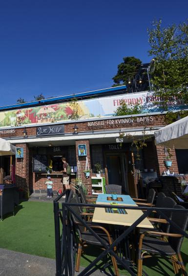 Underneath a brilliant blue sky, stands Pinehill Terrace's small, redbrick bar building and summer terrace, areas of seating under parasols either side and a green mat underneath them.