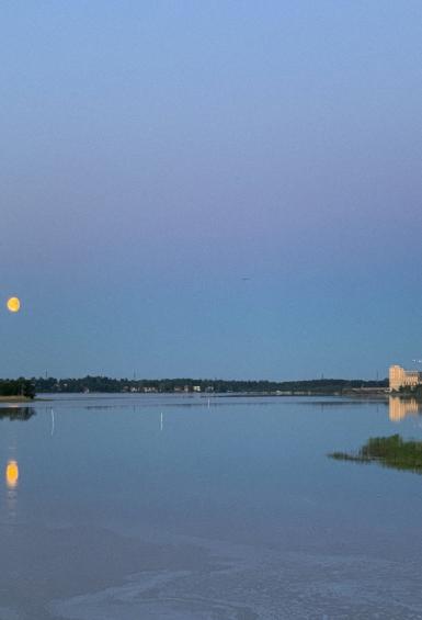 With a yellow moon against a deep blue sky at dusk, the bay next to Lammassaari stretches to shoreline on the horizon, where the tall buildings of Kalasatama standing on the right are lit by the setting sun.