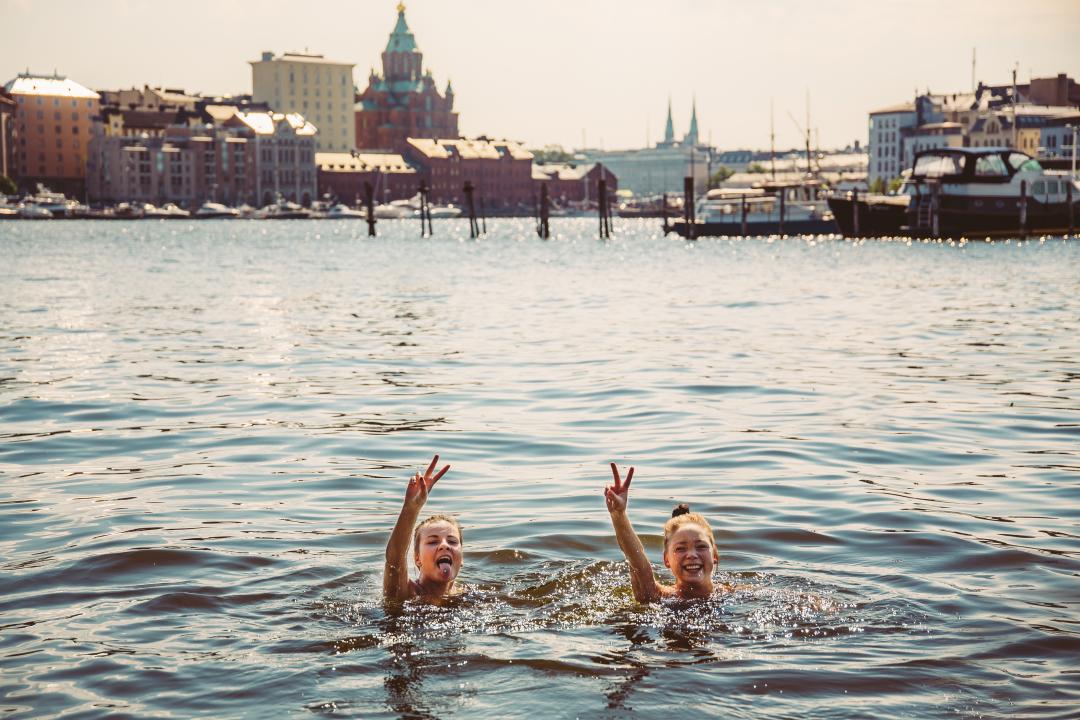 Two women swimming in Katajanokka bay on a sunny summer's day, stop to throw peace signs at the camera.