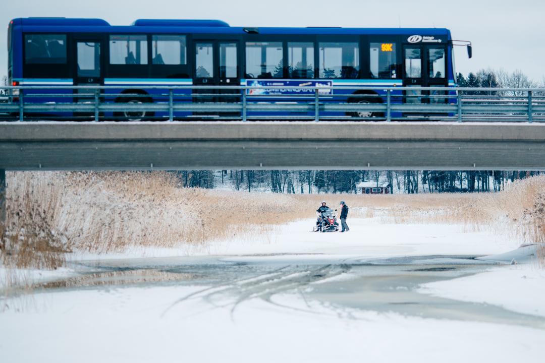 Viewed from the side, a bus crosses a bridge over a frozen river in winter. In the distance two men are chatting whilst standing on the river, one is sitting on a snow mobile.