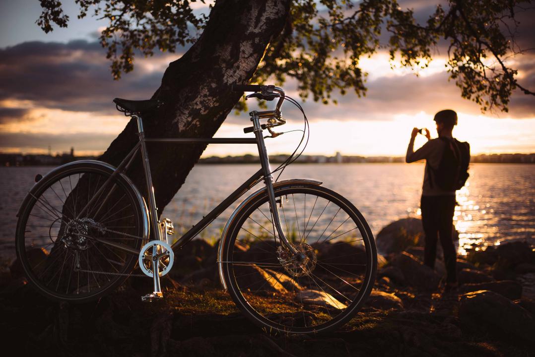 In the foreground a bike rests against a tree while a man stands on the shoreline in the background taking a photo of the sunset across the bay.