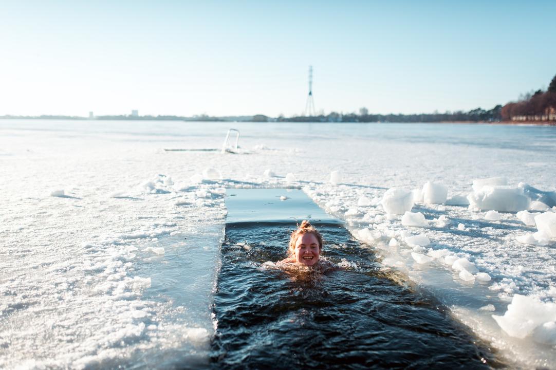 With the landscape of an ice covered sea dusted with snow stretching to the coast on the horizon, a smiling woman swims in a strip cut in the ice between trips to the sauna.