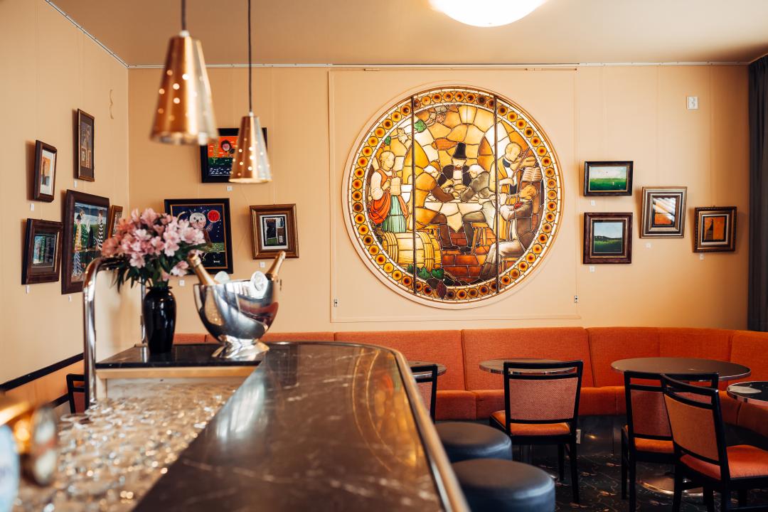 Running down the center of the photo away from the camera is a dark wood bar with dozens of empty glasses in a recess behind it, champagne bucket and a vase of pink flowers standing at the bar's end. The walls are salmon pink, with several small, framed paintings on the wall alongside a circular stained glass mural beneath which, small, round tables, chairs and an orange sofa sit.