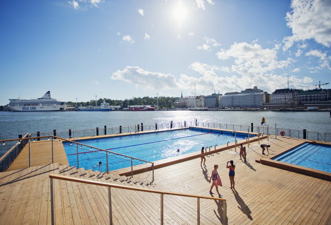 Standing at the top of some stairs leading down to Allas Sea Pool's main deck, people can be seen on the main deck walking to and from the main swimming pool, the harbour and Eteläranta shoreline in the distance. 