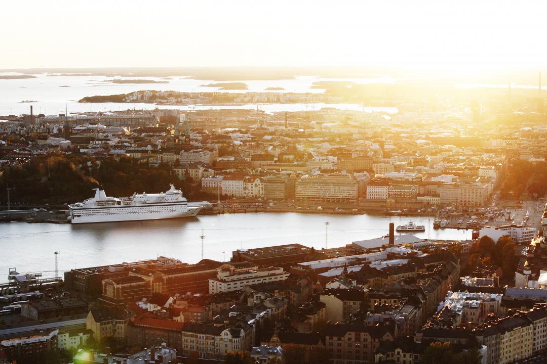 An aerial view looking over Katajanokka, Helsinki harbour and stretching into the distance, as seen from above Katajanokka. A ferry is at dock in the harbour, and the sunset shines golden and bright in the top right of the photo. 