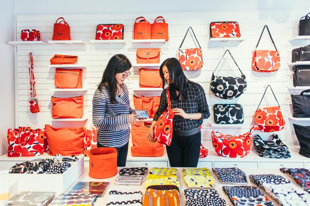 At a Marimekko store, wwo women stand in front of a handbag display that covers the wall whilst browsing Marimekko bags. A long table with a variety of Marimekko purses is in front of them. 