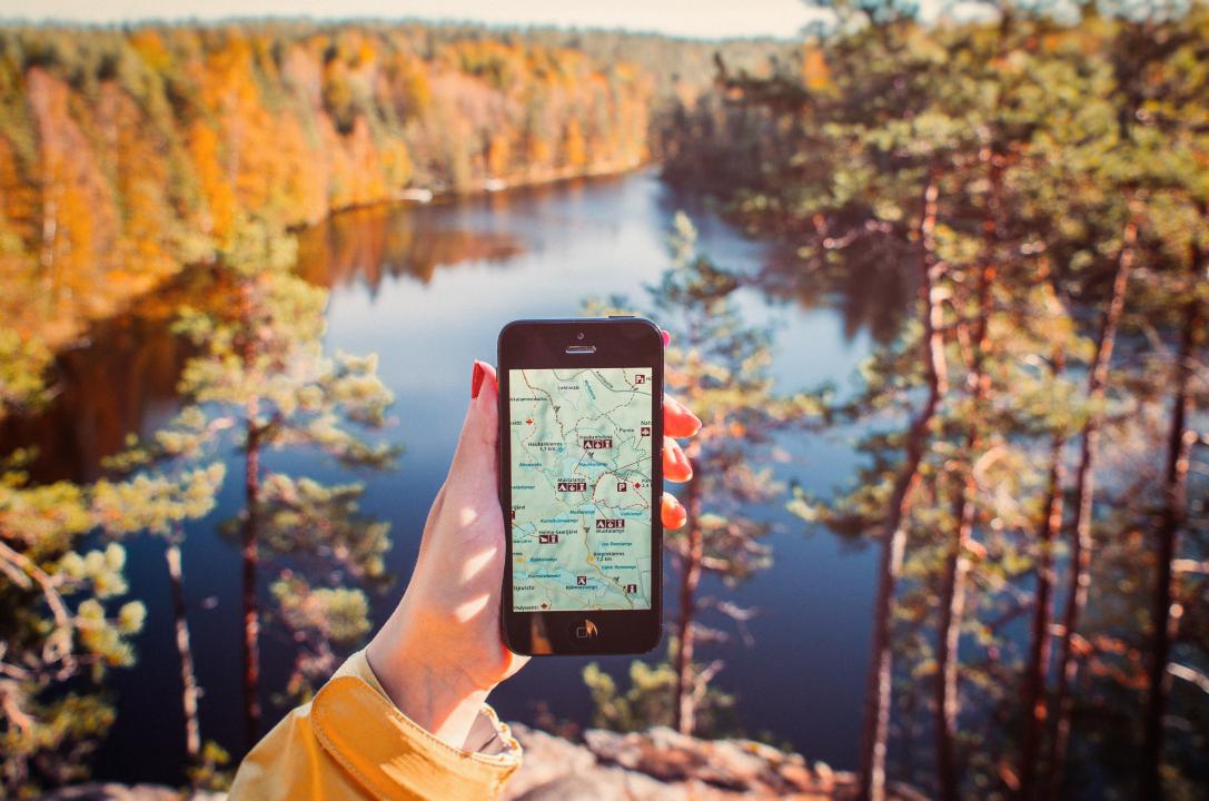 In the centre of the photo a hand is holding a mobile phone displaying a map of Nuuksio national park. In the background is a lake from the park viewed from above, surrounded by trees, some of which have become yellow in the autumn.