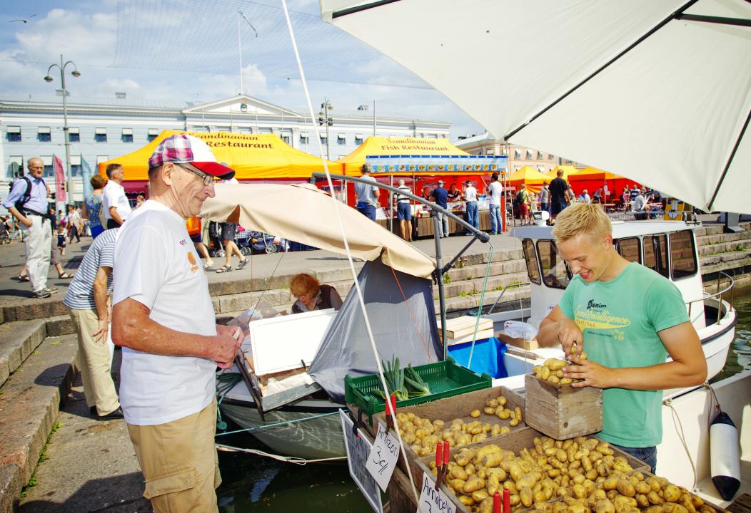 On the right a youn man sells new potatoes from the back of his boat to and older gentleman standing on the edge of helsinki Harbour. Helsinki Market Square's orange stall canopies can be seen in the background.