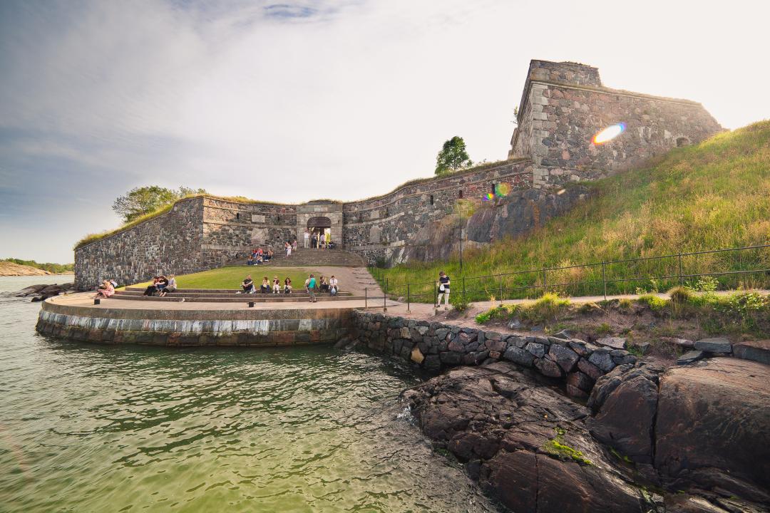 A view looking towards King's Gate on Suomenlinna where a few small groups of people are sitting on the steps and enjoying the sea view. A grassy bank rises up to the right of the photo.