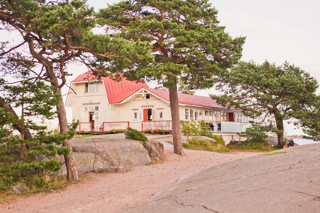 Raised on a rocky plateau behind three large trees, is a large, traditional wooden villa, an example of one of the many beautiful villas of Hanko.