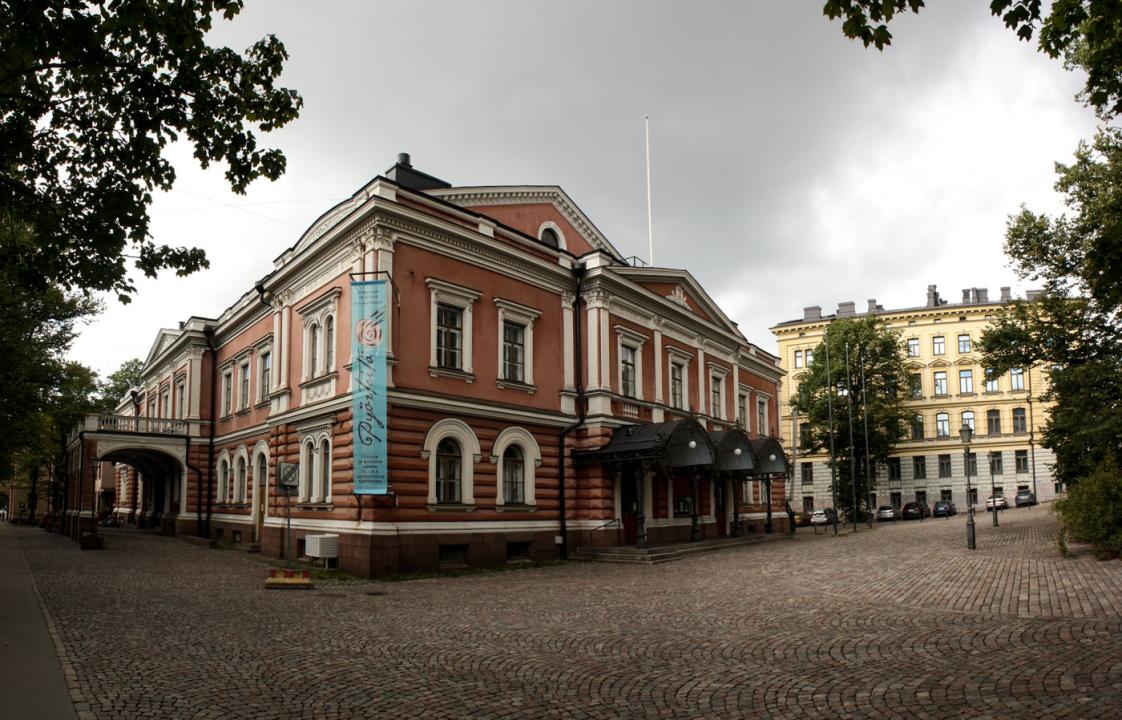 Ahead is the corner of the Alexander Theatre, the front facade facing the right and the length of the theatre leading to the left. Surrounding the theatre are cobblestone  pavements and a scattering of trees.