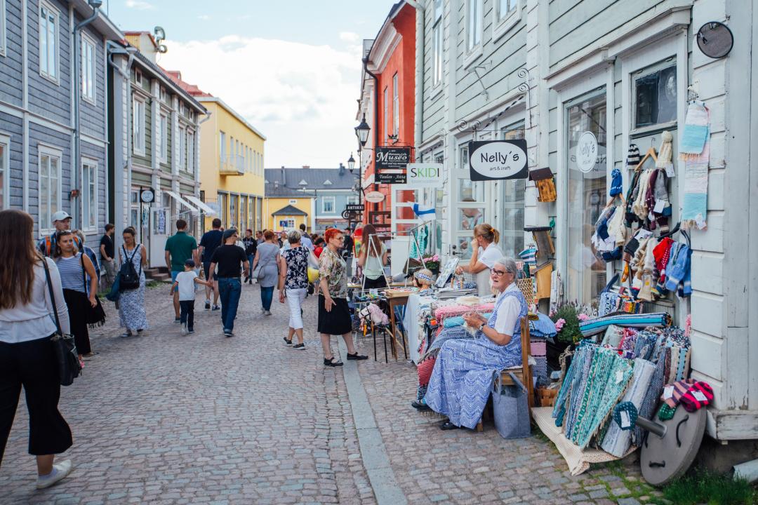 The narrow cobblestone streets of Porvoo's Old Town are lined with charming boutiques in historical wooden buildings of different colours. On the right two women are selling a variety of handcrafts outside their shops.