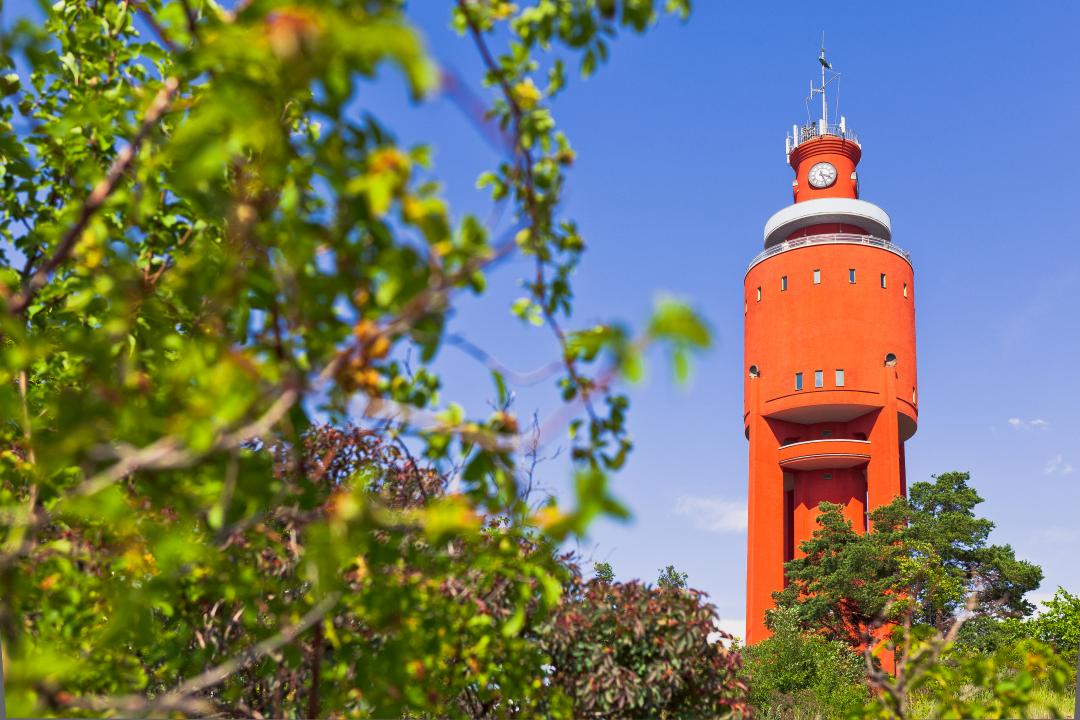 Viewed from behind trees in the left of the picture, Hanko’s terracotta orange water tower rises above the treeline against a blue sky.