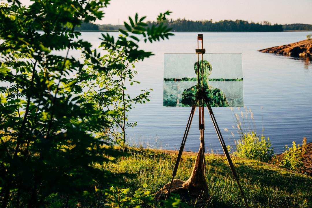 Next to a tall bush on a bankside at Kivinokka, a painting stands on a wooden easel, the sea stretching out to the opposite coastline on the horizon behind it.