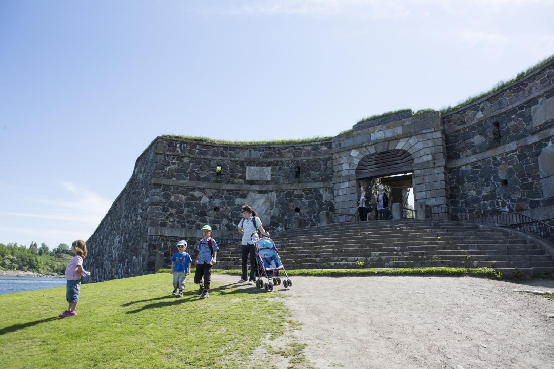 A woman stands with a pushchair and her three small children in front of King's Gate, Suomenlinna, on a bright and sunny day.