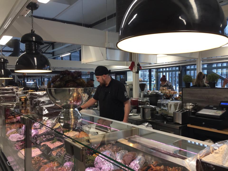 A man in black clothing serves a customer whilst standing behind a meat counter in a butcher's store at Hakaniemi's temporary market hall.