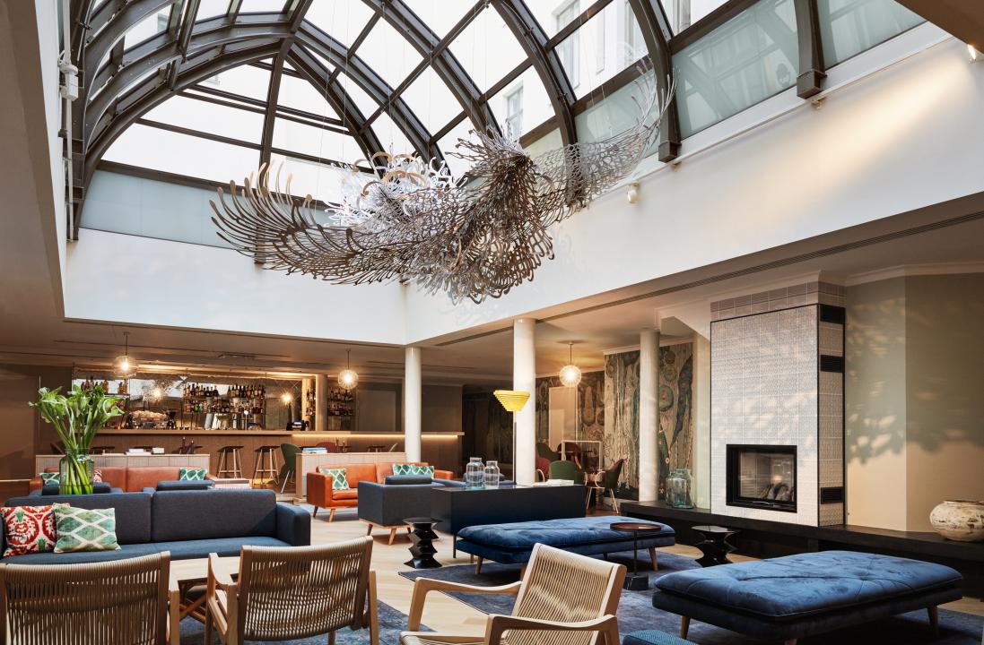 Hotel St.George Wintergarden, a large seated area filled with luxurious sofas and chairs underneath a skylight that stretches almost the whole length of the room. Pekka Jylhä's "Learning to Fly" sculpture hangs from the ceiling of the skylight..