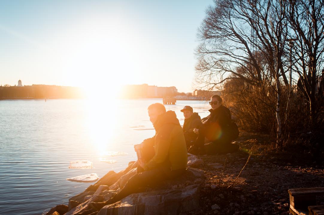 A group of three friends sitting on the shore of Sompasaari looking to the left, very strong sunshine shines across the sea,partially obscuring the Helsinki cityscape in the distance.