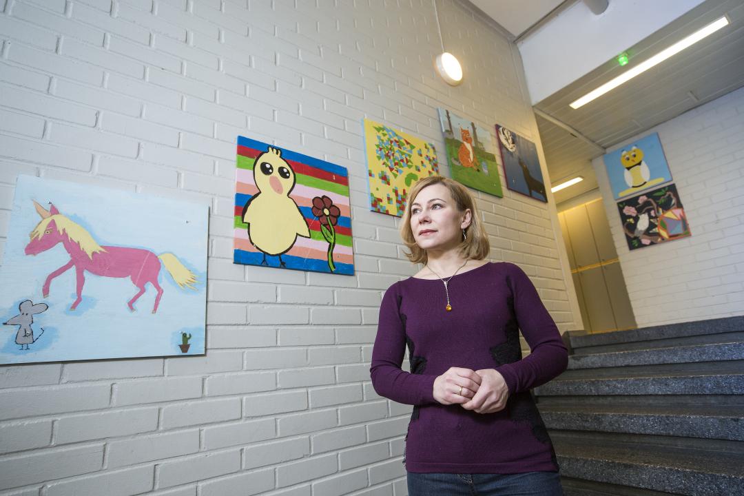 Looking towards the left, Jelena Nerman is standing in front of a small set of steps in a corridor, colourful kids paintings on the wall behind her.