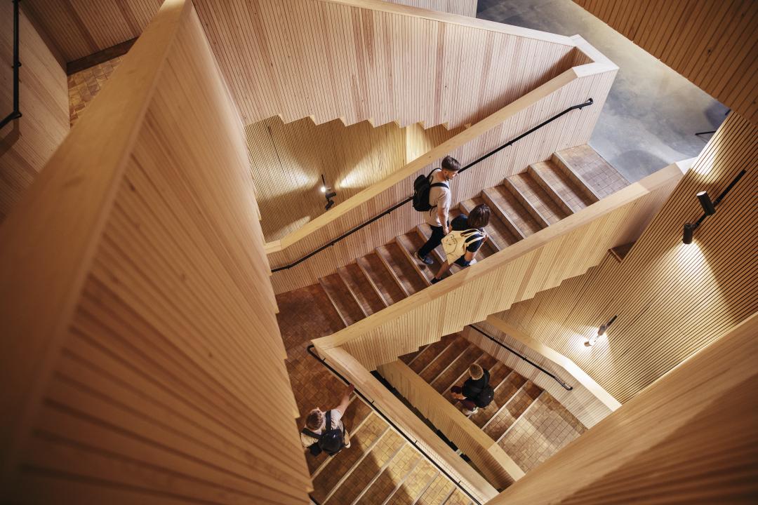A view looking down on 4 people using the stairwell of Helsinki University Think Corner. The whole interior is made of light natural wood and the staircase zig-zags across the space of the stairwell as it descends. 